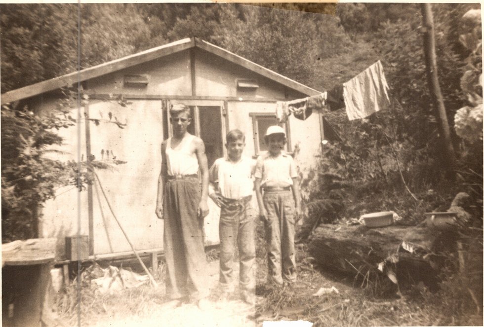 Three children standing outside a small corrugated-iron shed on a bush block, a clothesline strung beside it